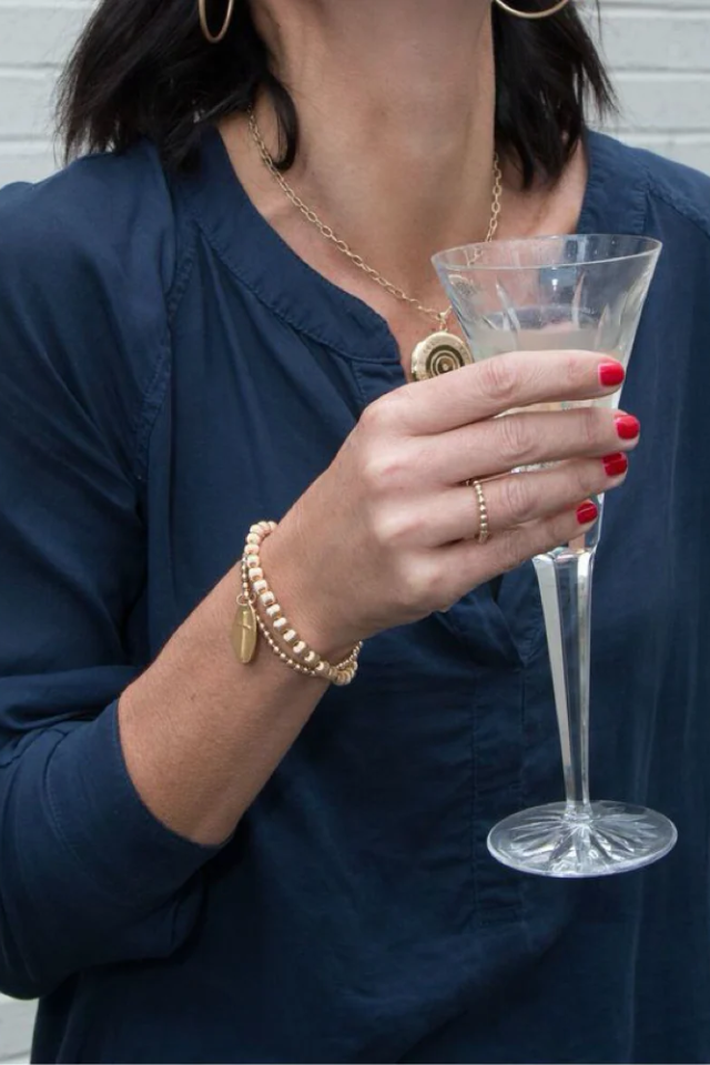 Person holding a clear glass with a dark background. Gold beaded bracelet on a white background. enewton. 14k gold, tarnish free. low price. 